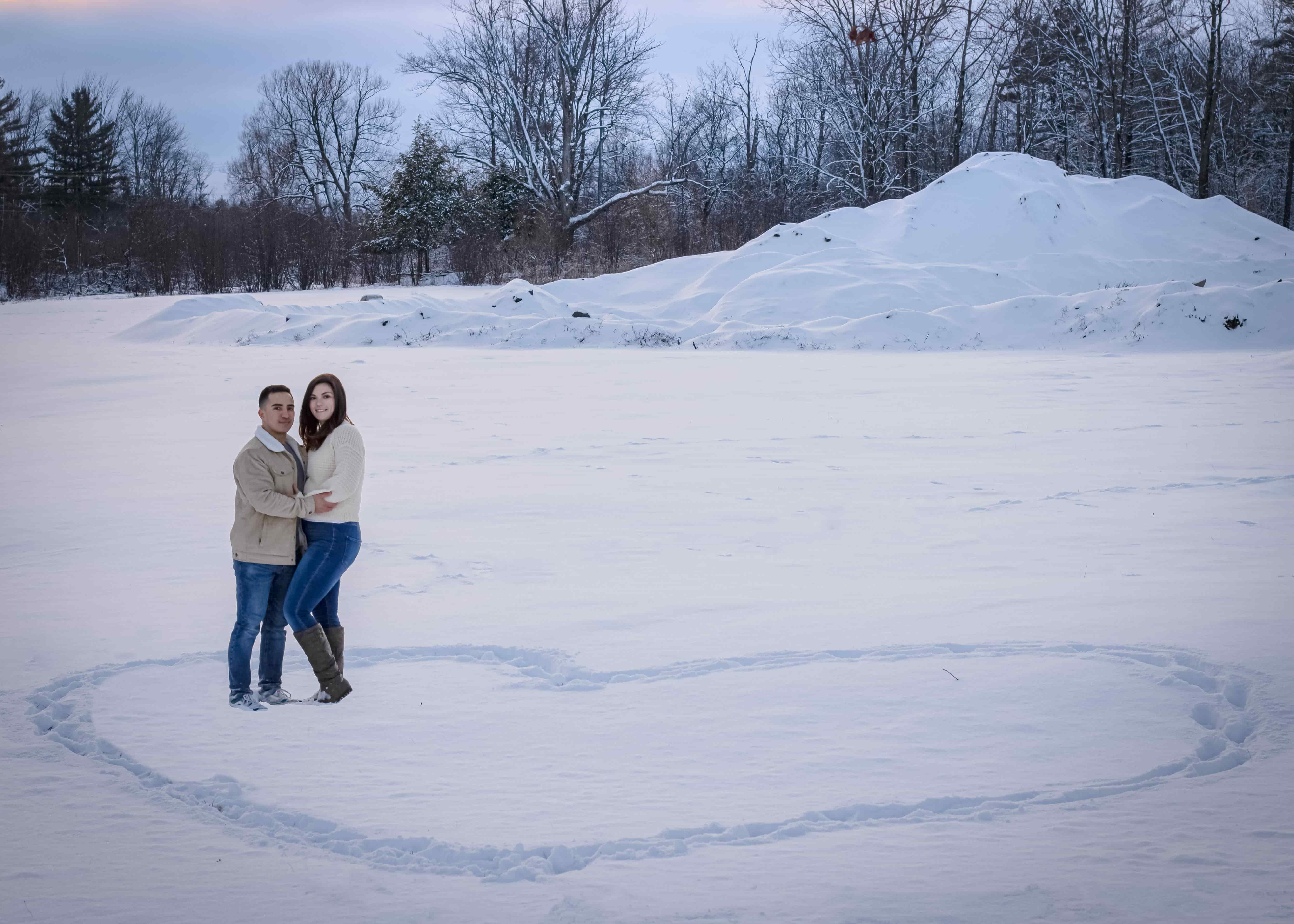 A man and woman in an open field of wood and snow- standing inside of a heart drawn in the snow
